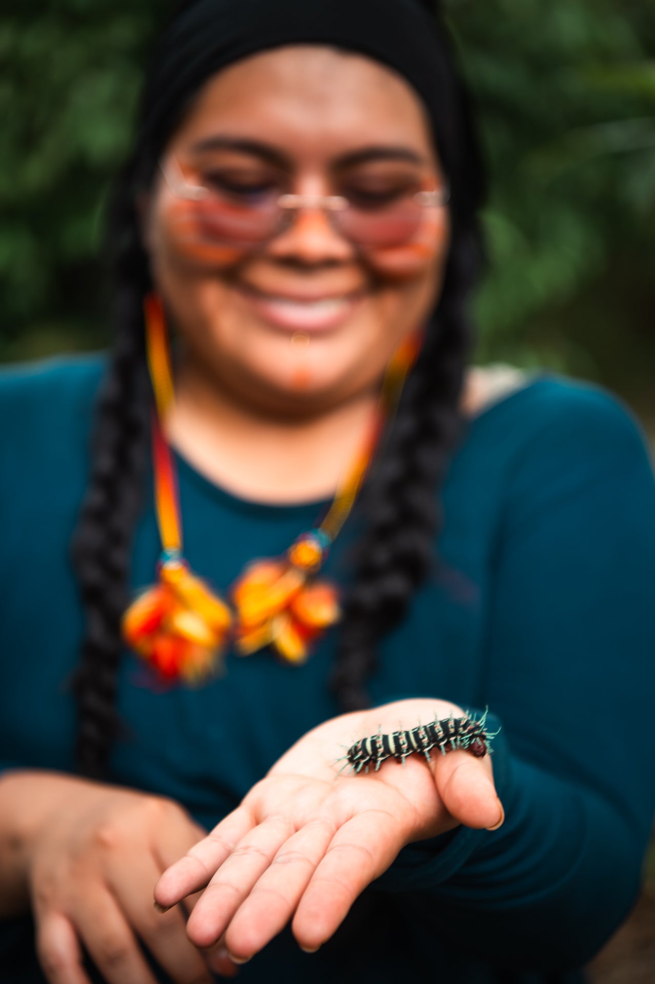 A smiling woman wearing a black headscarf and a teal long-sleeve top holds her hand out with a small black and green caterpillar resting on her palm. She is wearing a colorful orange and yellow beaded necklace and glasses, with a soft-focus green outdoor background.