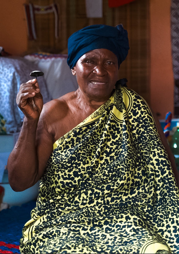 A smiling woman seated indoors wearing a dark blue headwrap and a yellow-and-black patterned draped garment, holding a small mushroom in one hand.