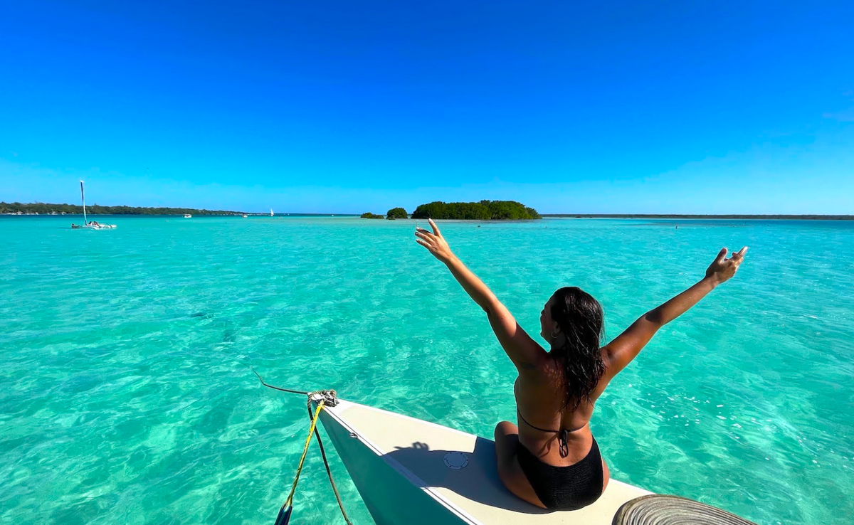 A woman in a black bikini sits on the bow of a small boat, facing the turquoise laguna with her arms raised, under a clear blue sky.
