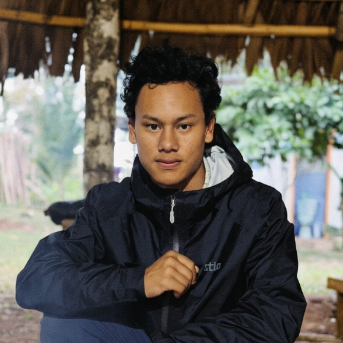 A man wearing a black hooded jacket, navy pants, and muddy boots crouches under a thatched roof structure in an outdoor rural setting.